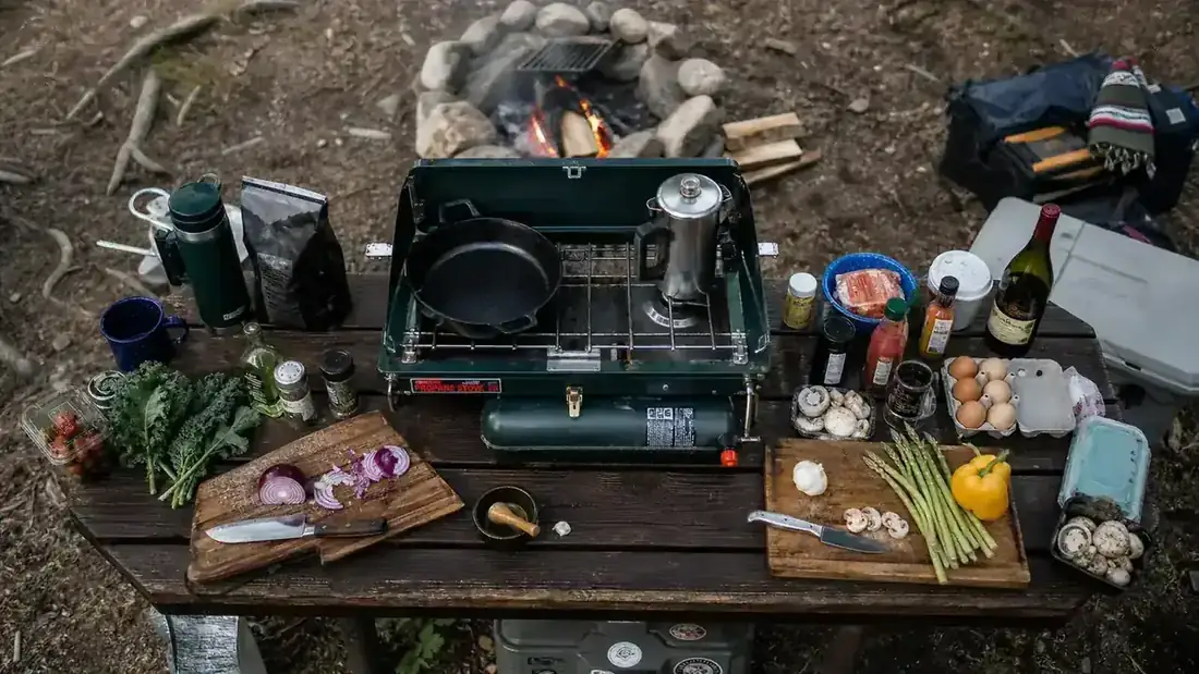 Campsite cook standing at a two-burner stove flipping pancakes while a second pan holds sausage links on the other burner