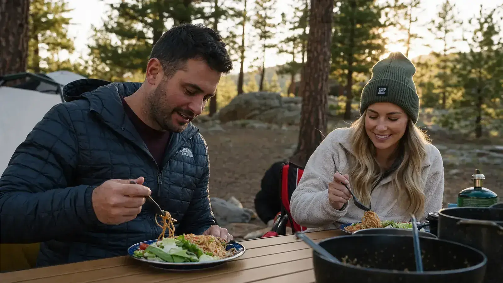 A completed camp meal of scrambled eggs, bacon, and toast plated on a camp plate next to a mug of coffee at a picnic table